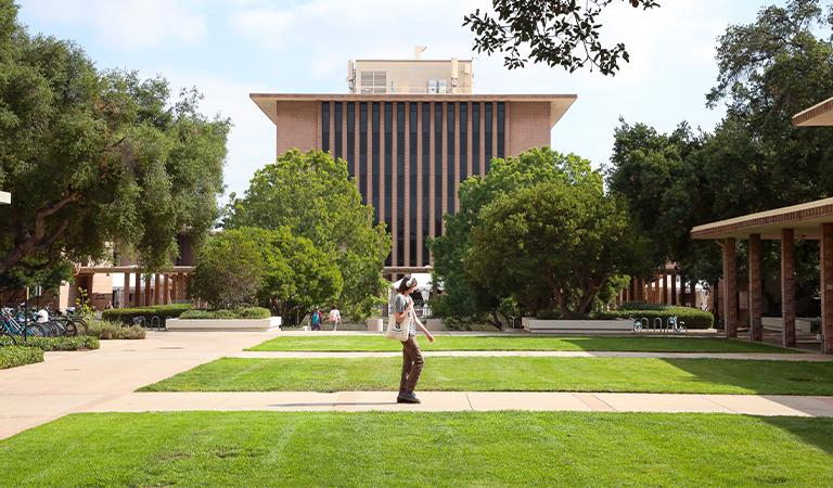 A student holding a tote bag and wearing headphones walks across campus.